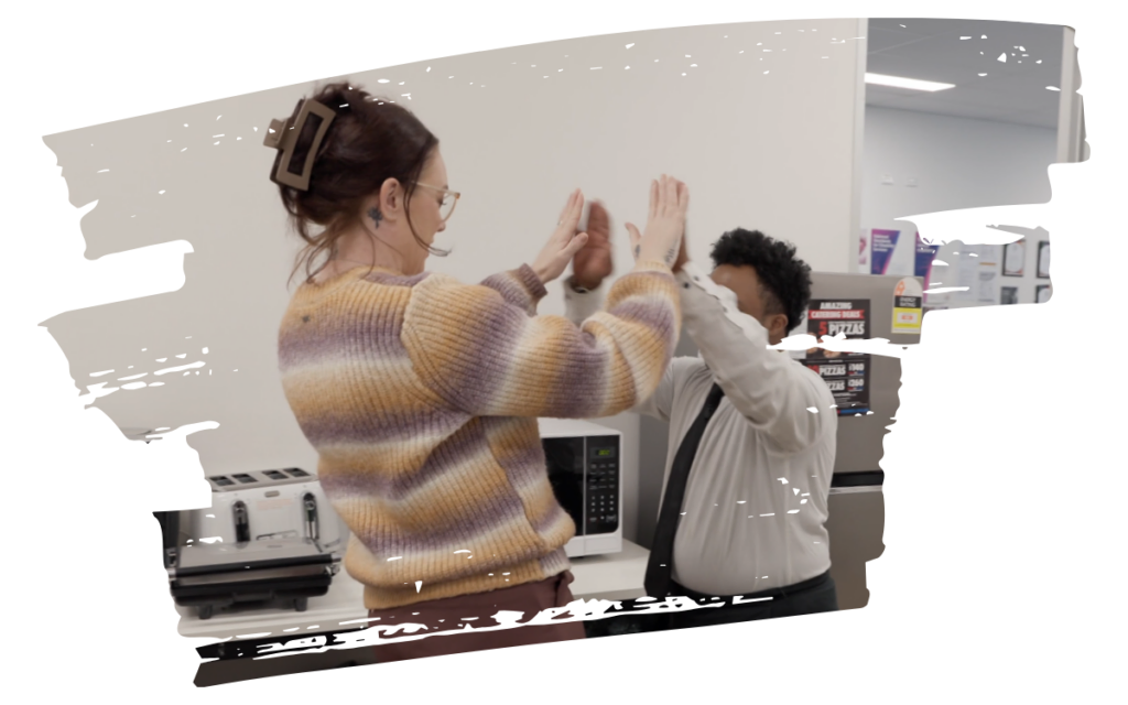 Two coworkers share a high-five in a office kitchenette area beside a microwave and toaster.