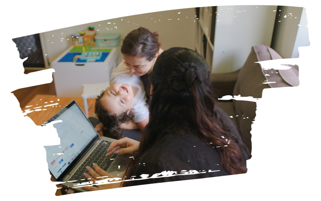 Child lying on a table with a big smile, while an adult leans over and another person works on a laptop nearby in a cozy room.