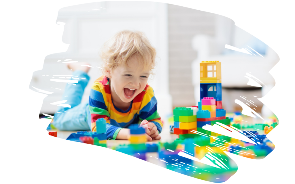 Young child lying on the floor, smiling, while playing with colorful building blocks in a bright playroom.