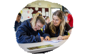 Two students sit at a classroom table, writing on papers while a smiling woman assists nearby in a busy classroom.