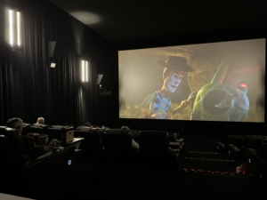 Dark movie theater with a large screen showing Woody and Rex from Toy Story; audience seated in the foreground.
