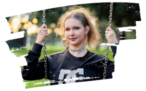 Young woman with blonde hair sits on a swing in a sunny park, wearing a black DC sweatshirt and a sparkling choker.