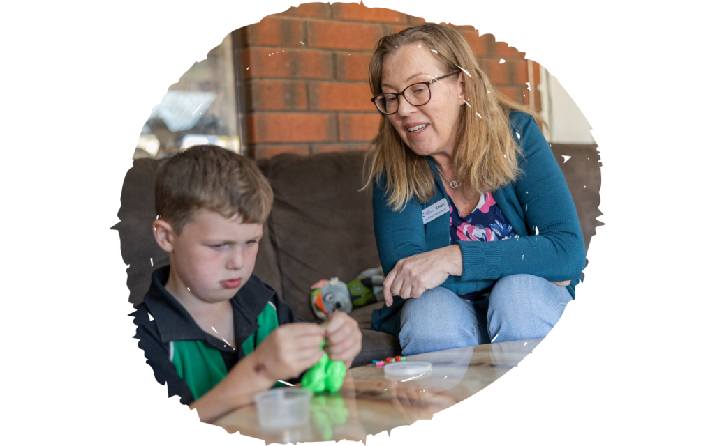 Woman with glasses helps a boy with a hands-on craft at a low table in a cozy room with a brick wall behind them.