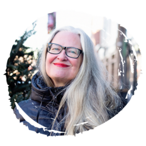 Portrait of a smiling woman with long gray hair and black glasses, wearing a dark puffer jacket outdoors.