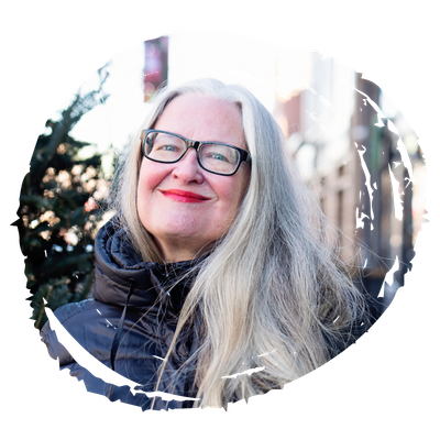 Portrait of a smiling woman with long gray hair and black glasses, wearing a dark puffer jacket outdoors.